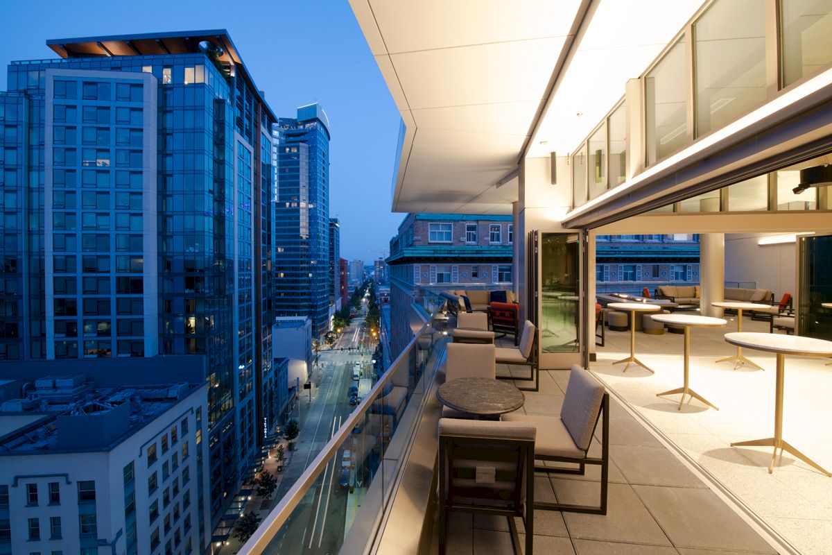 A modern cityscape at dusk with tall buildings and an illuminated, stylish outdoor balcony area featuring tables and chairs.