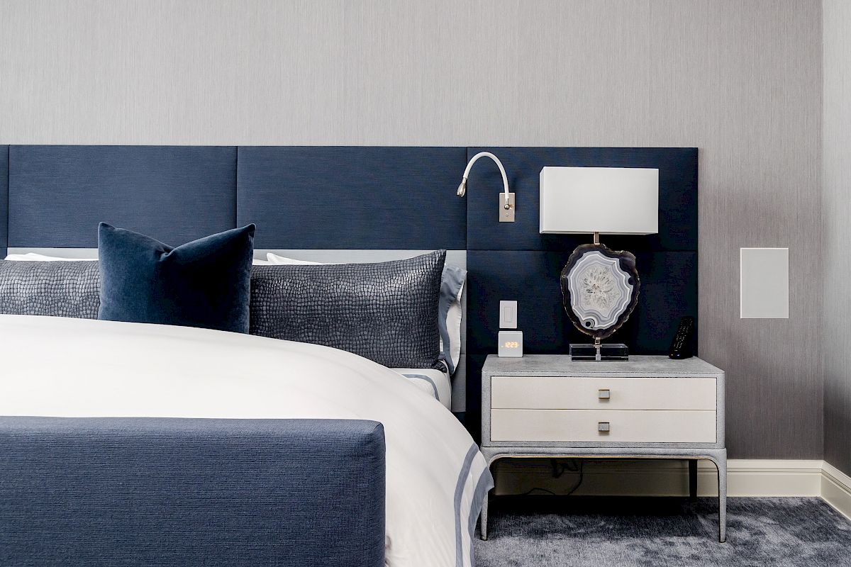 A modern bedroom features a navy headboard, white and navy bedding, a nightstand with a lamp and decorative item, set against a gray wall.