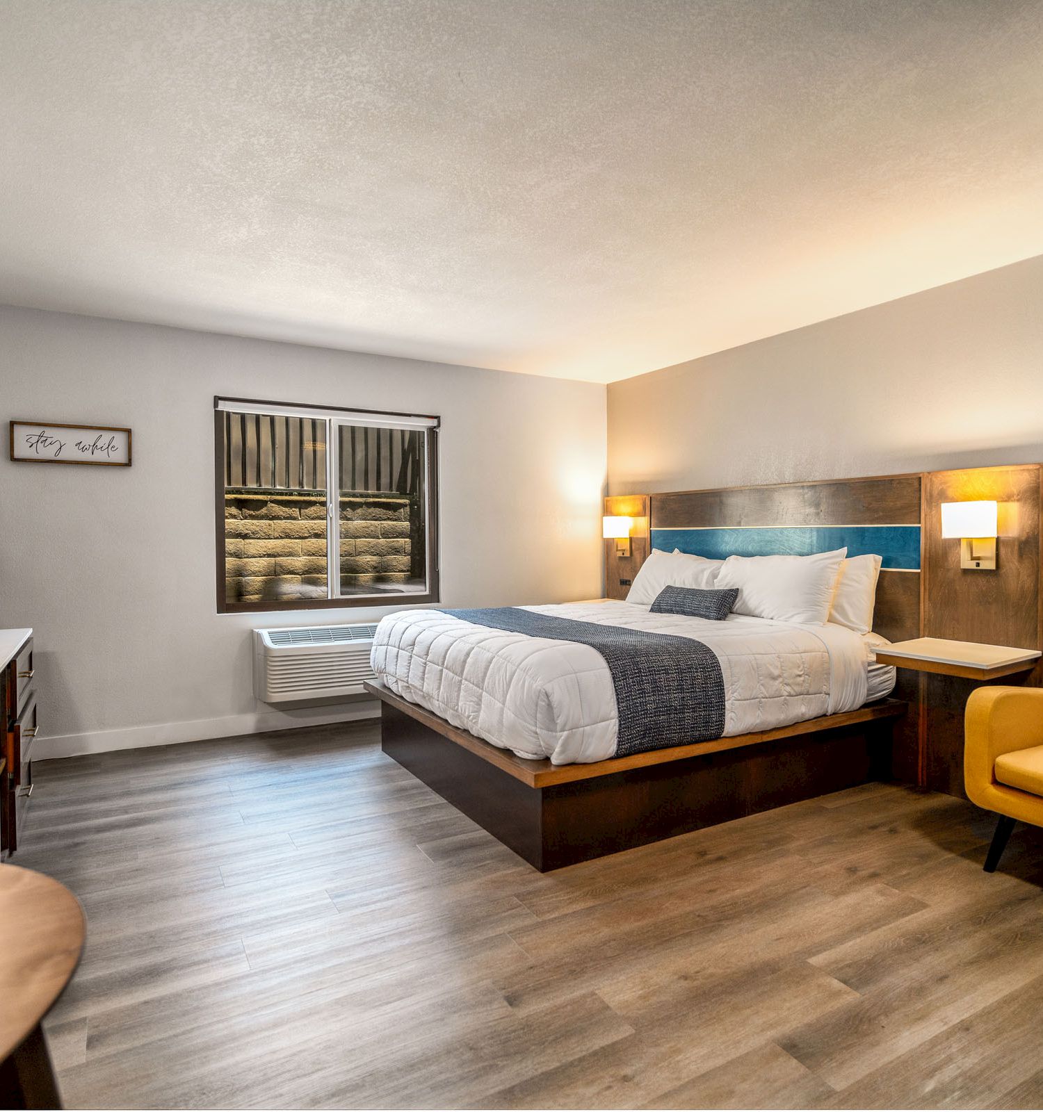 A modern hotel room features a double bed, yellow armchair, wooden furniture, TV, window, and wall-mounted reading lamps on either side of the bed.