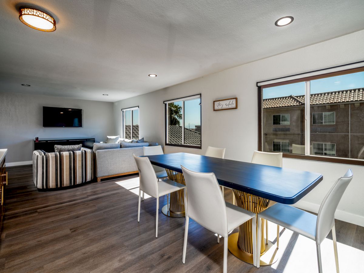 A modern living and dining space with a rectangular blue table, white chairs, a striped sofa, TV, and large windows letting in natural light.