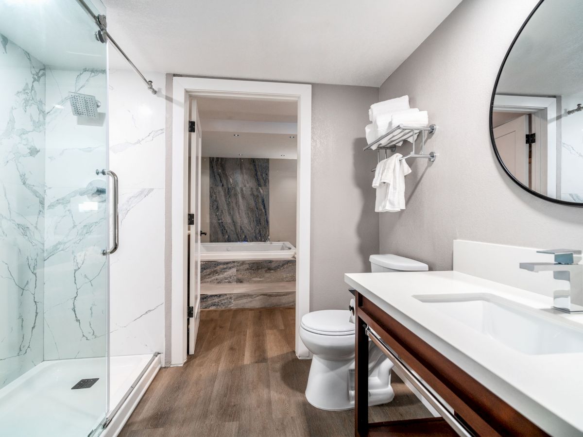 Modern bathroom with a glass shower, white sink, round mirror, toilet, and towels on a rack. Visible bathtub in the adjacent room.