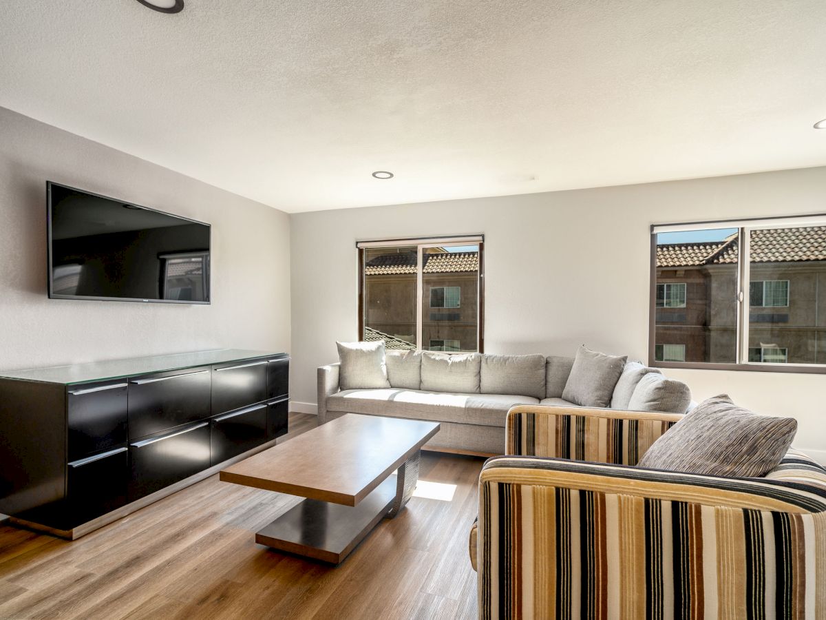 A modern living room with a wall-mounted TV, a long sofa, a striped armchair, a wooden coffee table, and two windows letting in natural light.