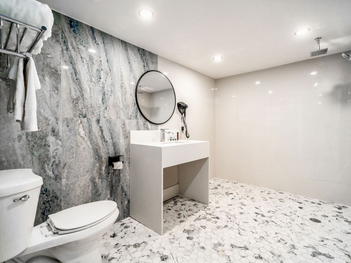 A modern bathroom with a toilet, vanity with a round mirror, and a shower area. The floor features hexagonal tiles with a mix of light and dark tones.