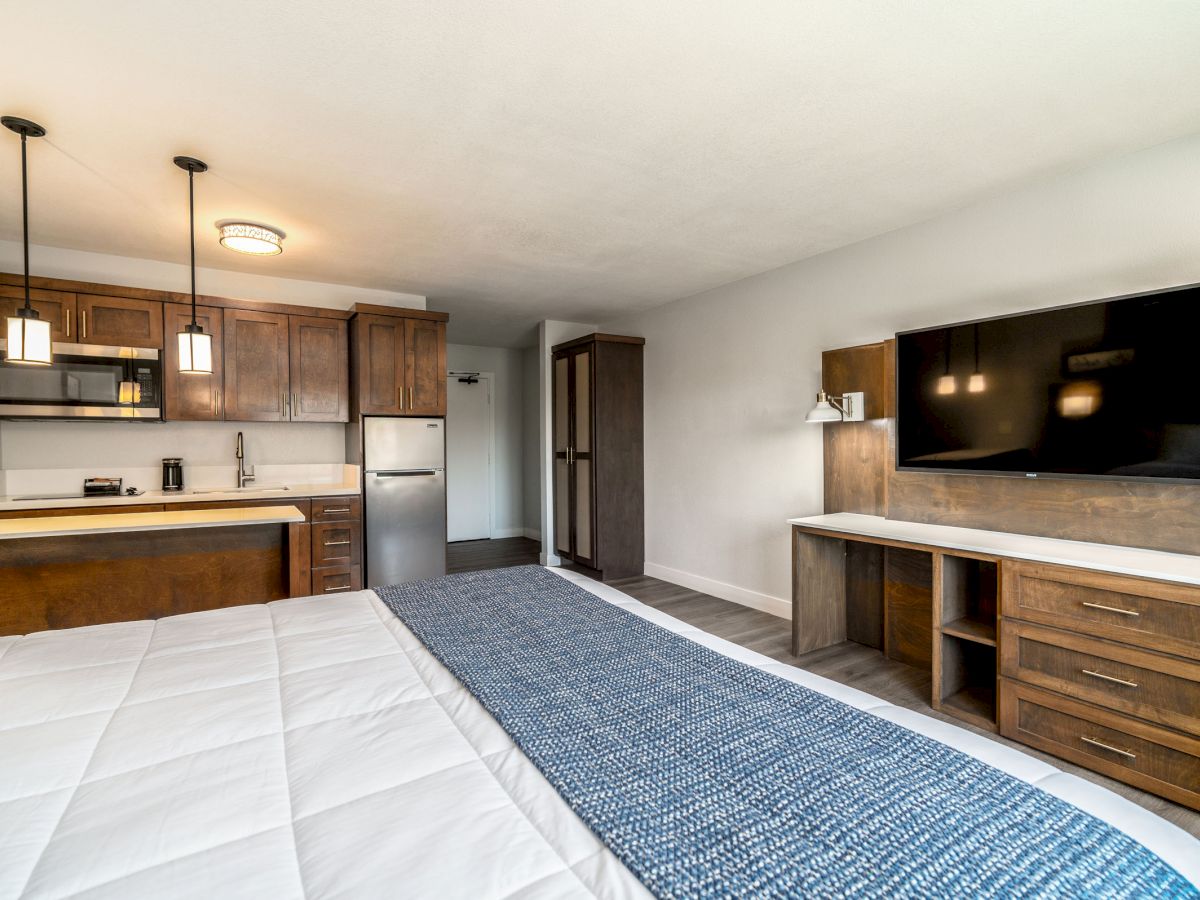 A hotel room with a double bed, a kitchenette, a large flat-screen TV, and wooden furnishings, including cabinets and a desk, ending the sentence.