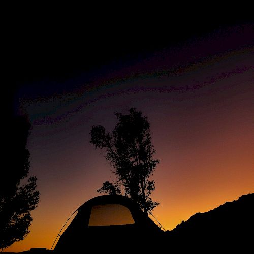 A silhouetted tent and trees against a colorful sunset sky with hues of orange, purple, and black.