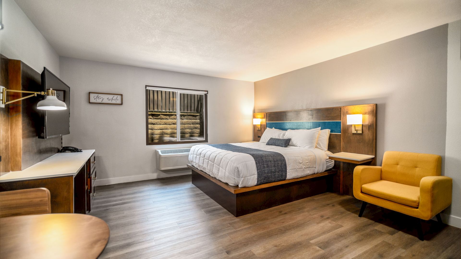 This image shows a modern hotel room with a large bed, yellow armchair, desk, wall-mounted TV, and window, all in a clean, contemporary style.