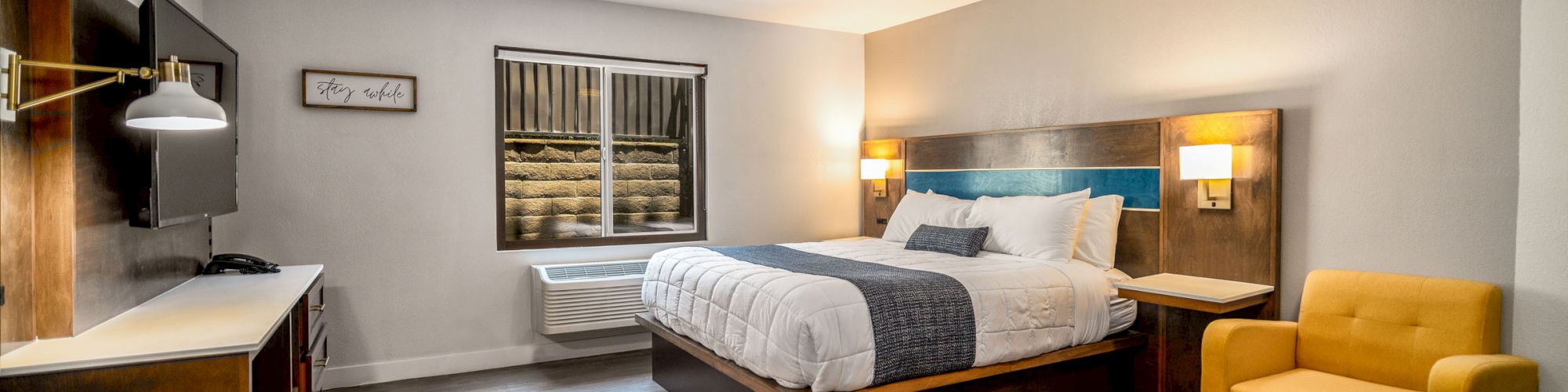 This image shows a modern hotel room with a large bed, yellow armchair, desk, wall-mounted TV, and window, all in a clean, contemporary style.