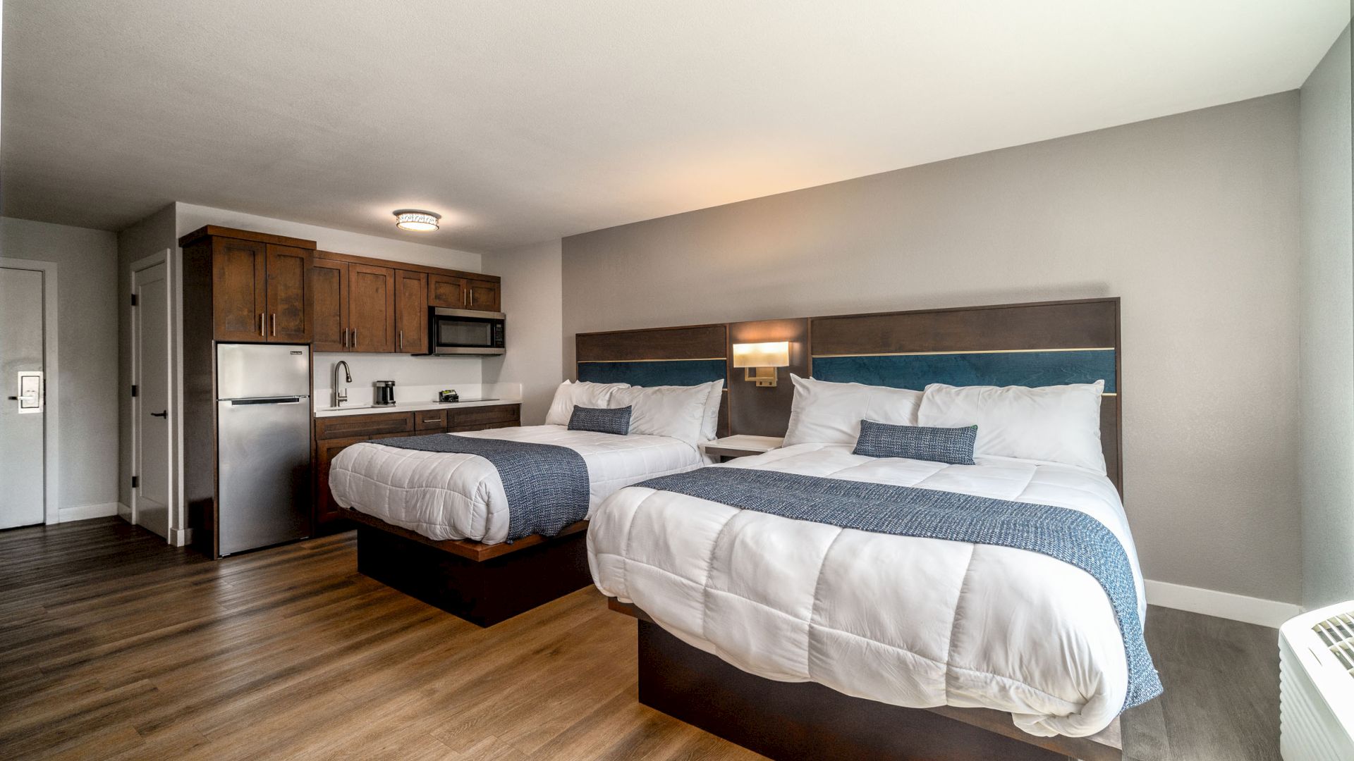 A modern hotel room with two large beds, a small kitchenette, wood flooring, and a minimalistic design.
