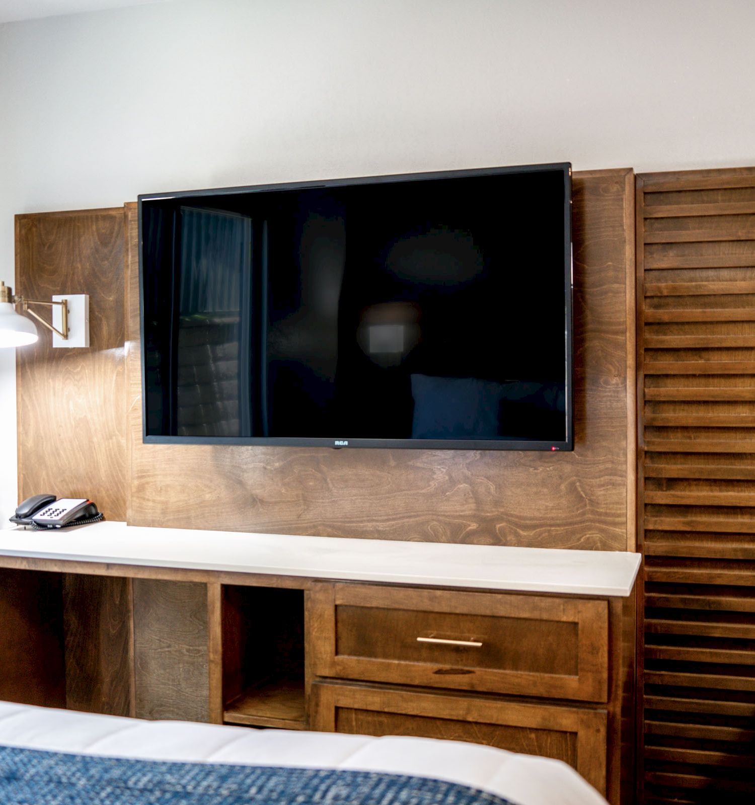 The image shows a hotel room with a wall-mounted TV, a wooden desk with drawers, a lamp, a telephone, and part of a bed. The wall has artwork reading 