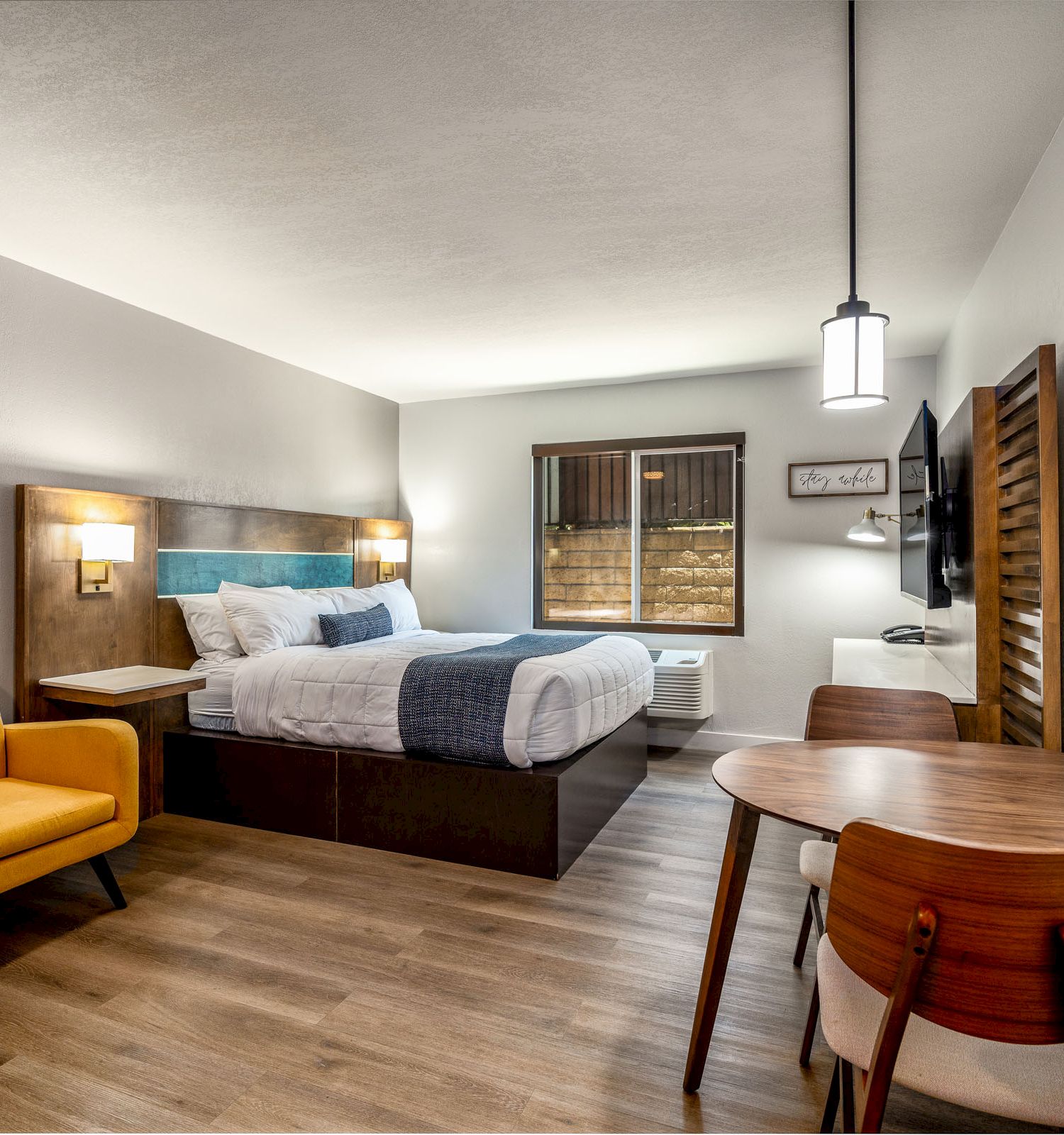 A modern hotel room features a bed, a yellow armchair, a wooden table with two chairs, a flat-screen TV on the wall, and a window with wooden blinds.