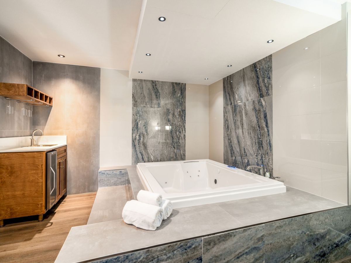 The image shows a modern bathroom featuring a large built-in bathtub with folded towels on the edge. There is a small sink and countertop area.