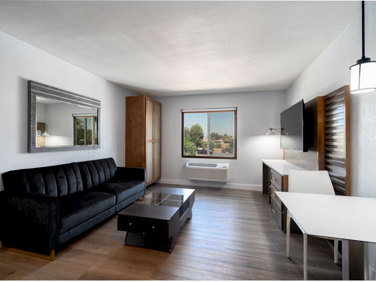 A modern living space with a black couch, coffee table, wall-mounted TV, window, mirror, and white table and chairs, with wood flooring.