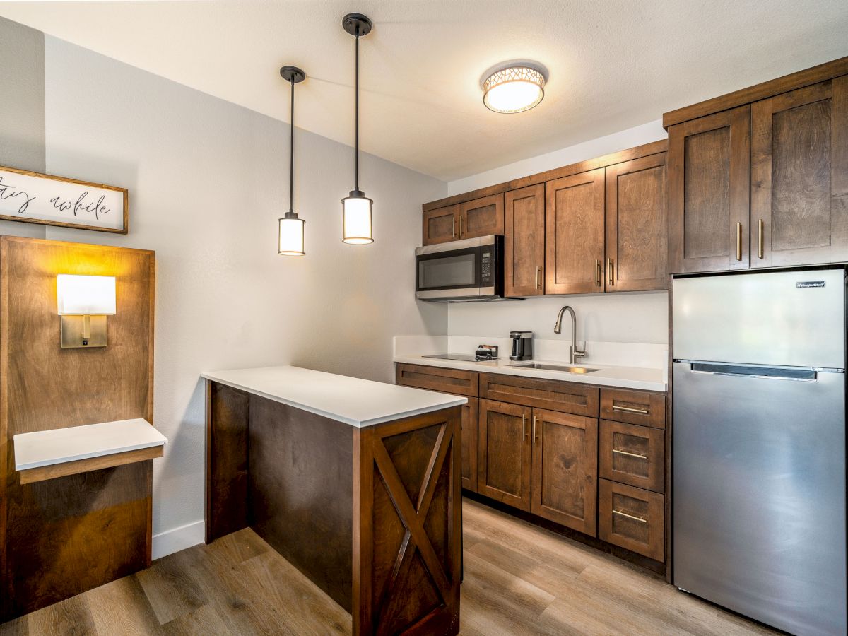 A modern kitchen with brown cabinets, a small island, pendant lights, a microwave, a fridge, and a wall sign reading 