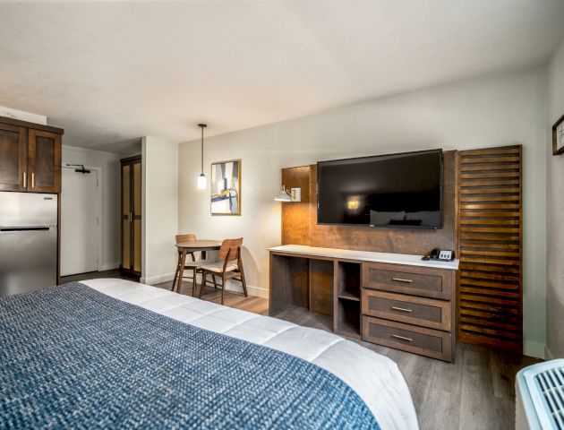 A cozy hotel room with a bed, a wall-mounted TV, a small dining table, a desk, a refrigerator, and wooden furnishings, creating a comfortable ambiance.