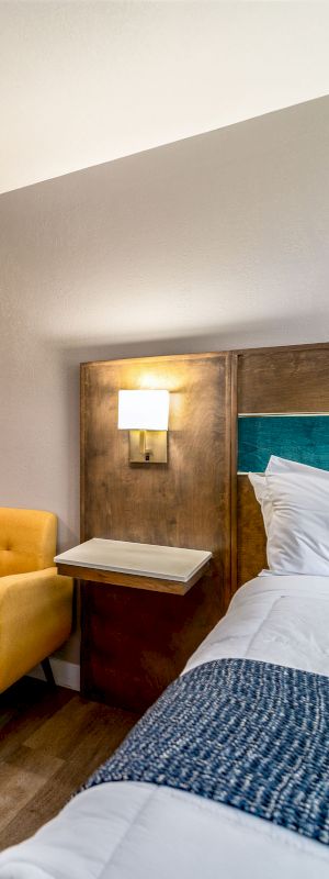 A hotel room with a kitchenette, a yellow chair, and a bed with blue and white bedding is shown in the image.