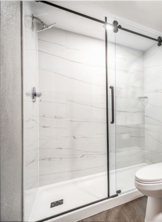 This image shows a modern bathroom with a shower, a toilet, a sink with a large mirror, and shelves holding towels. The shower has a glass door.