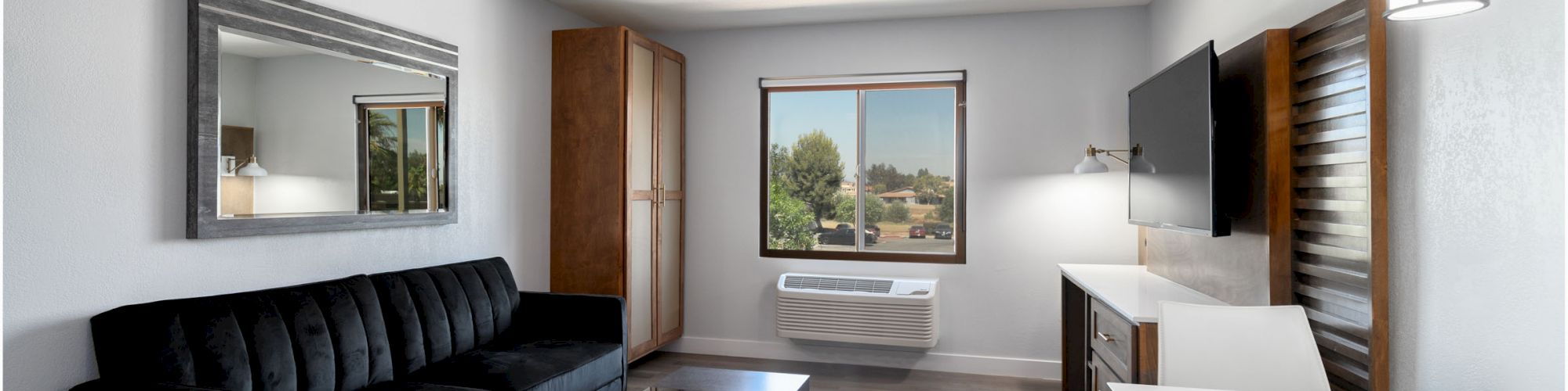 A modern living room with a black couch, coffee table, wall mirror, wardrobe, window view, air conditioning unit, TV, desk, and pendant light.