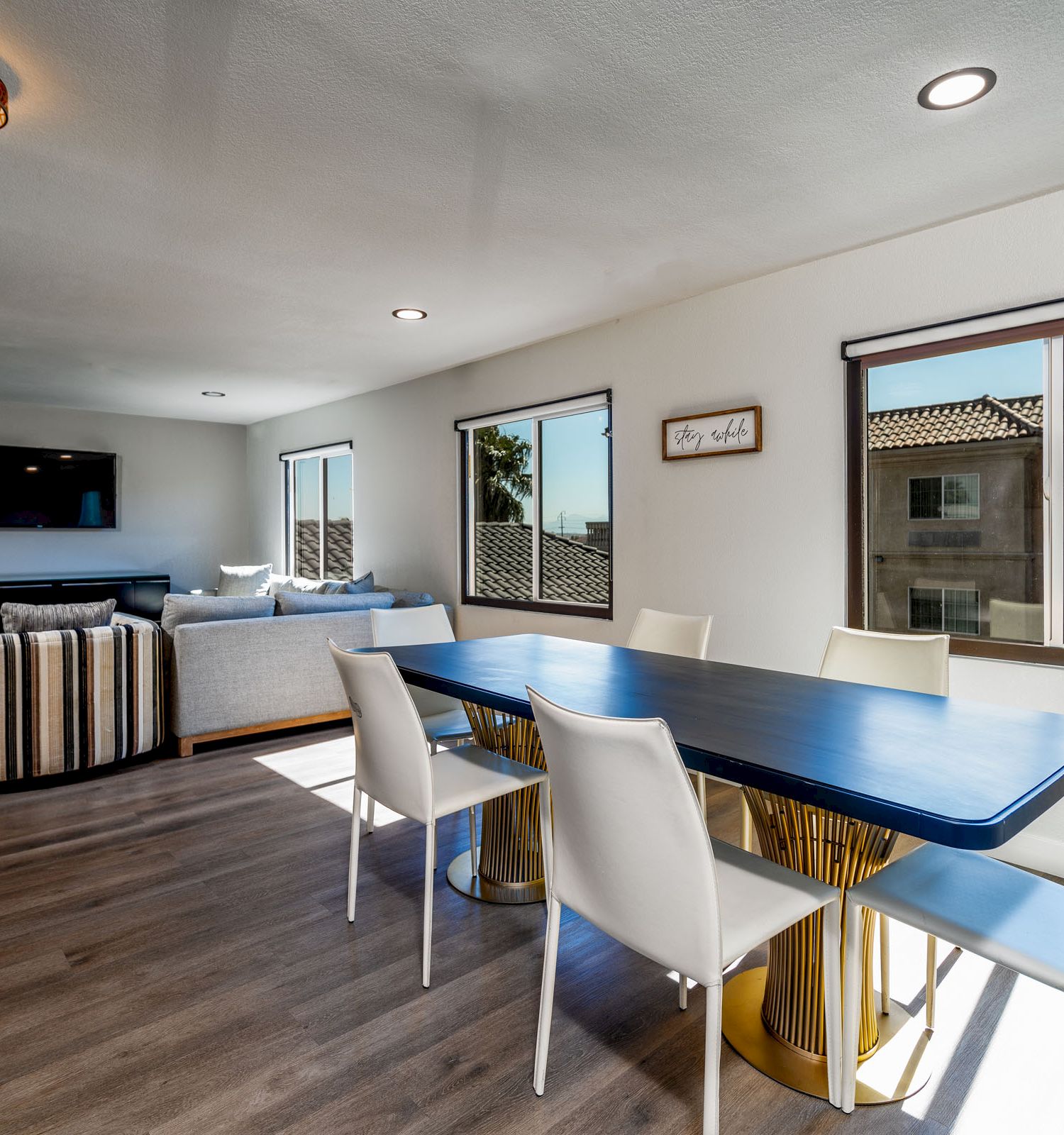 A modern dining area with white chairs, a blue table, and large windows. A living room with a TV and sofas is visible in the background.