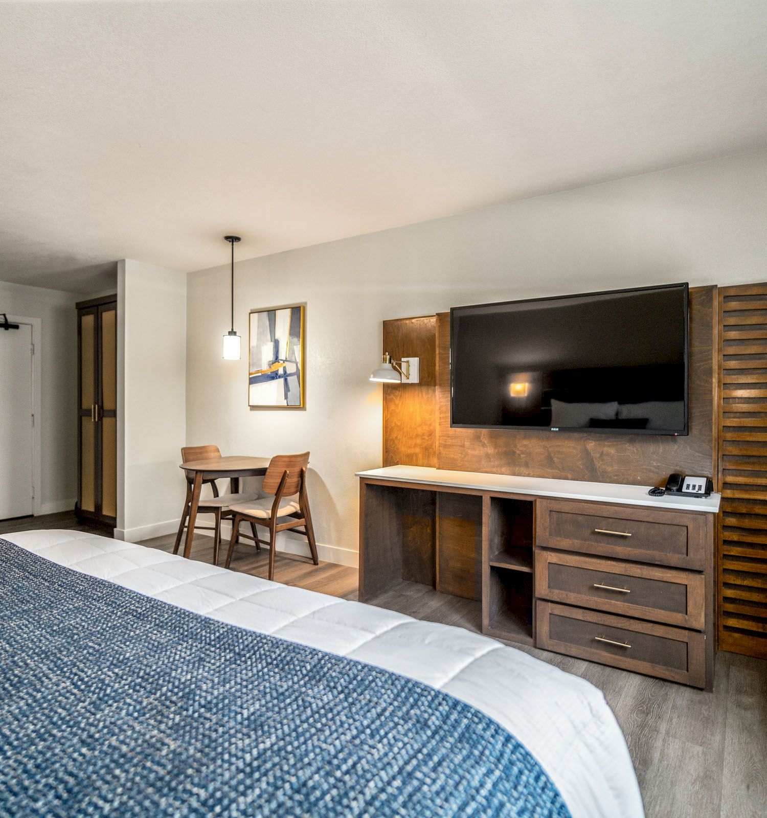 This image shows a modern hotel room with a bed, TV, desk, and chairs. There is also a kitchenette area with a fridge.