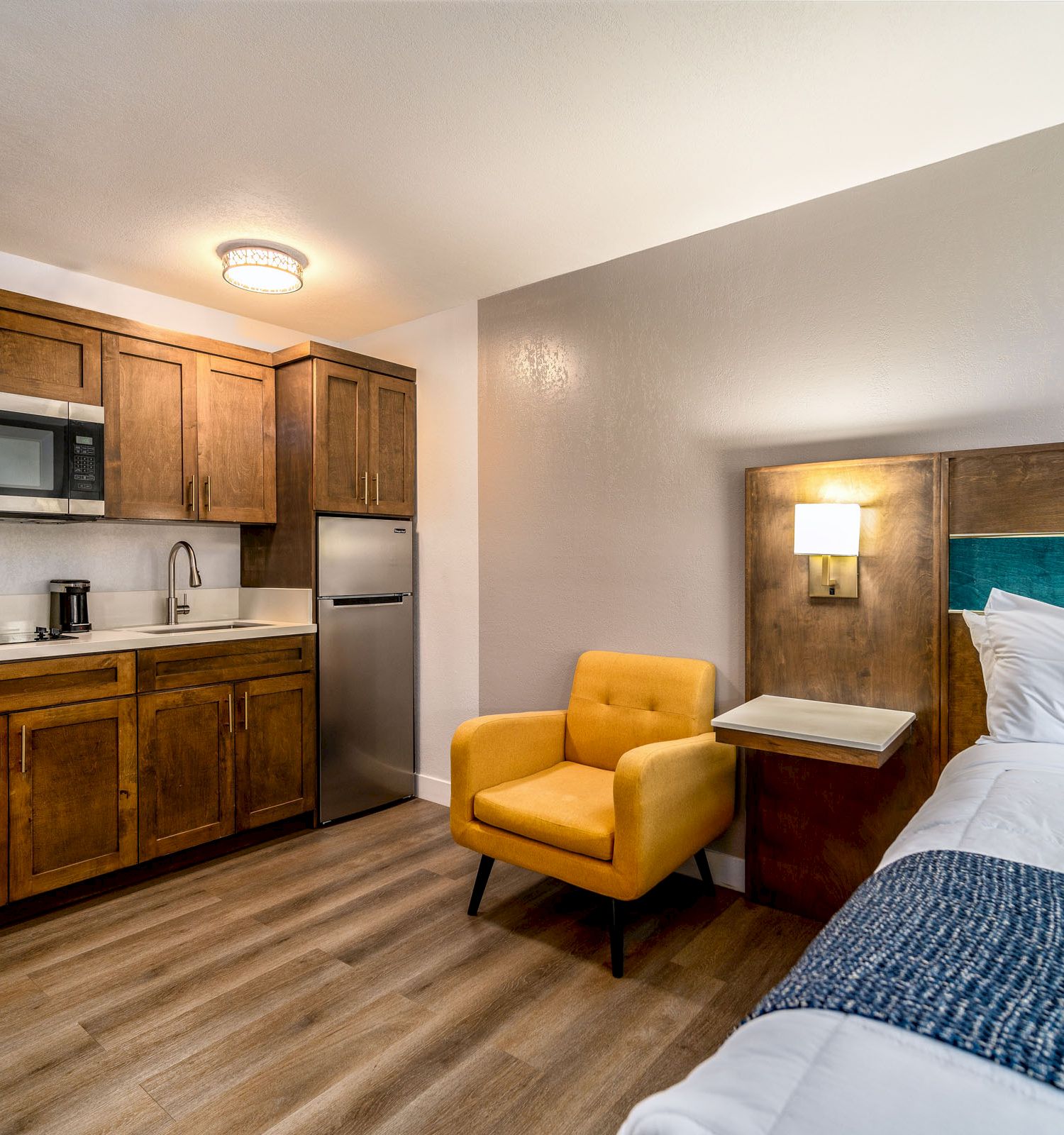 A cozy hotel room with a kitchenette, featuring a microwave, sink, and mini-fridge, a yellow armchair, and a neatly made bed with blue accents.