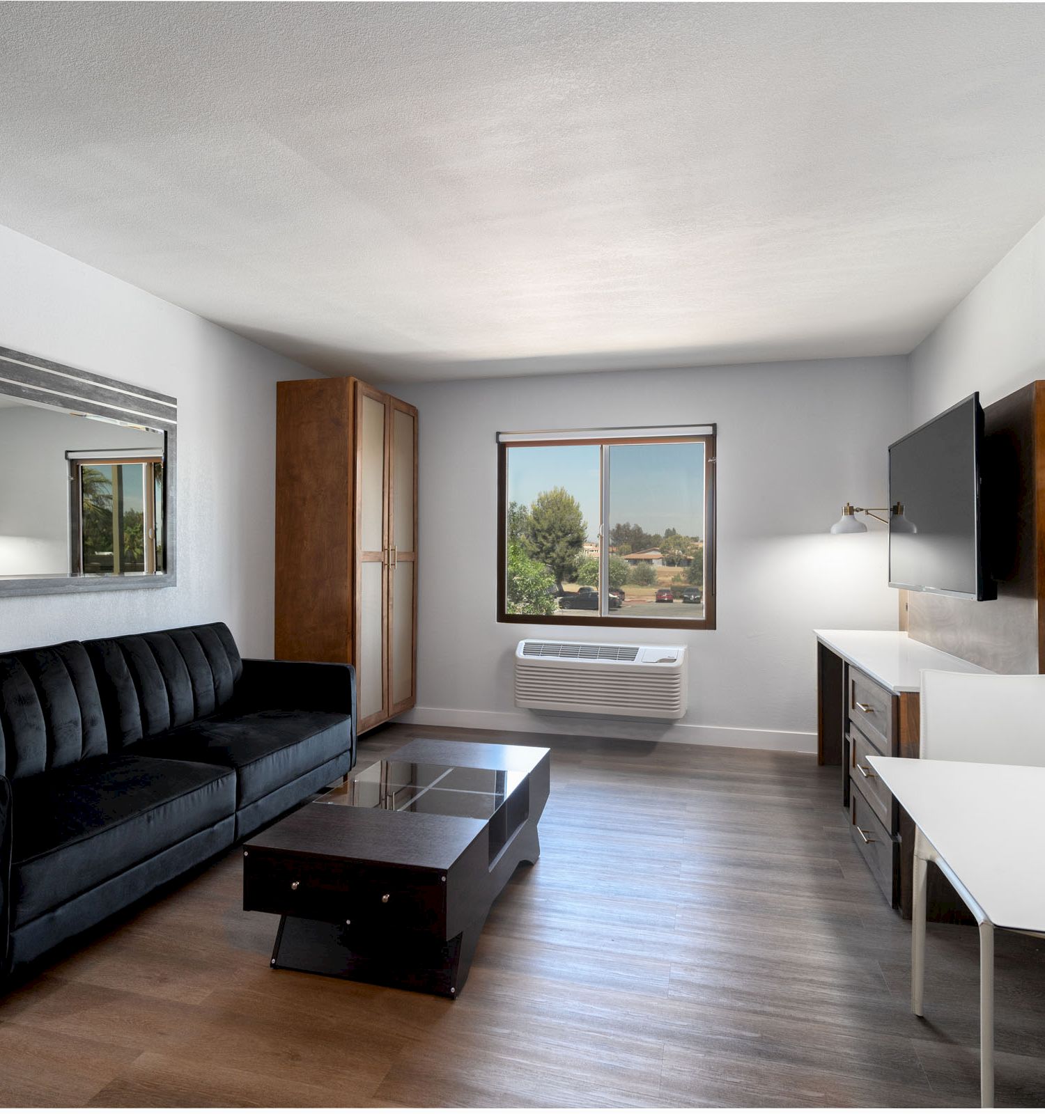 The image shows a modern living room with a black sofa, coffee table, flat-screen TV, wall mirror, window, desk, chairs, and wooden flooring.