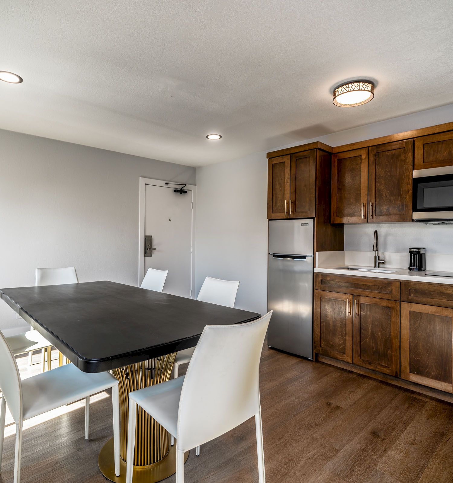 Small kitchen with wooden cabinets, a microwave, and a mini-fridge, alongside a dining area with a black table and white chairs.