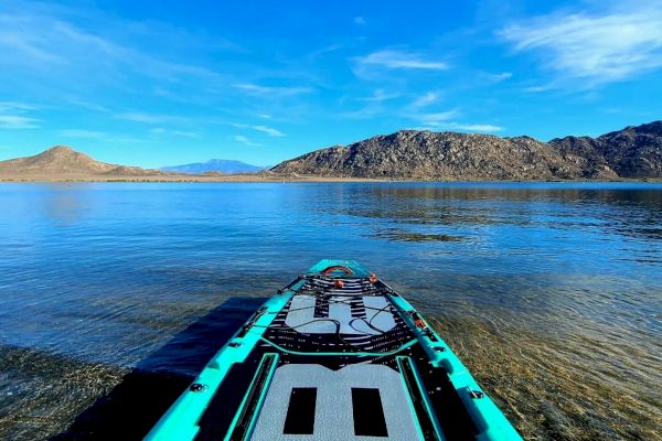 A paddleboard on a clear lake with mountains in the background under a bright blue sky, viewed from the board's deck.