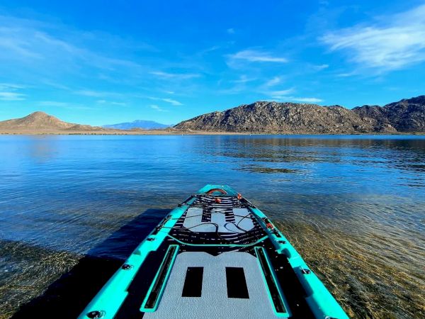 A paddleboard on a clear lake with mountains in the background under a bright blue sky, viewed from the board's deck.