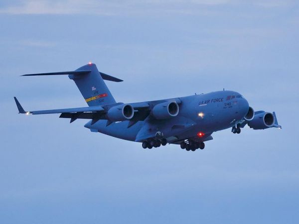 The image shows a military transport aircraft with the text "AIR FORCE" visible on its side, flying against a backdrop of a clear sky.