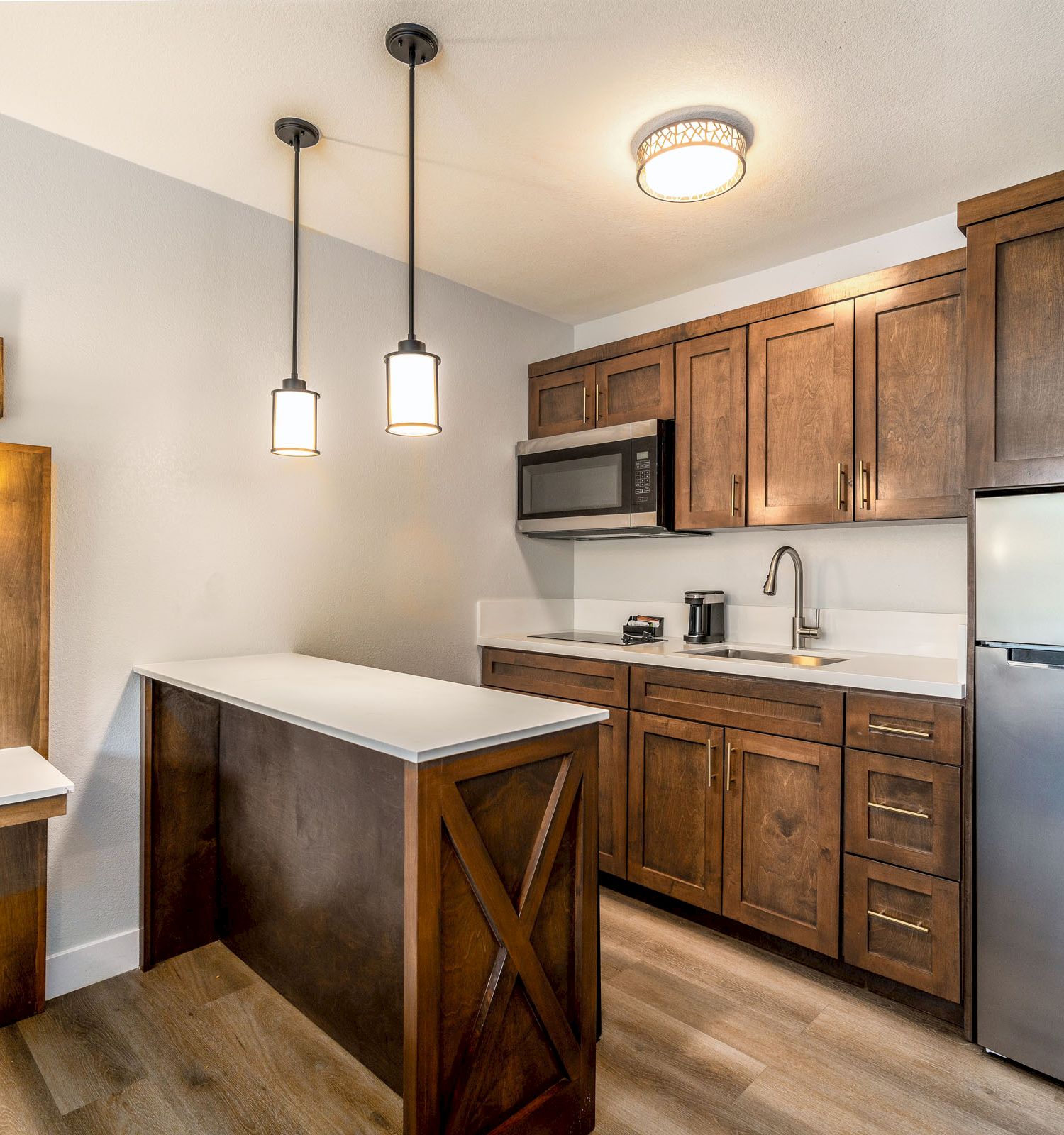 This image shows a small, modern kitchen with wooden cabinets, a fridge, a microwave, and pendant lights above a compact island.