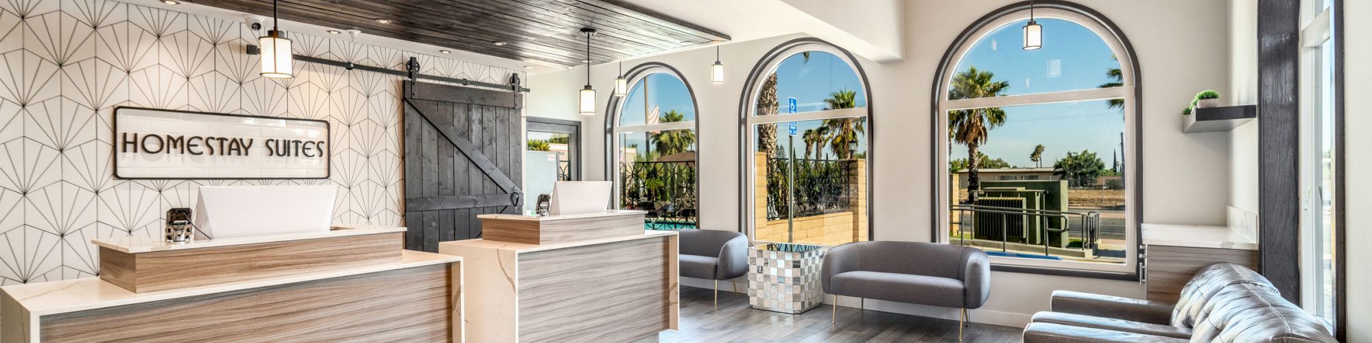 This image shows a modern hotel lobby with minimalist furniture, a reception desk, and large arched windows offering a view of palm trees outside.