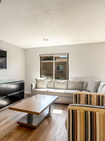 A living room with a gray sofa, striped armchair, wooden coffee table, wall-mounted TV, and two windows letting in natural light.