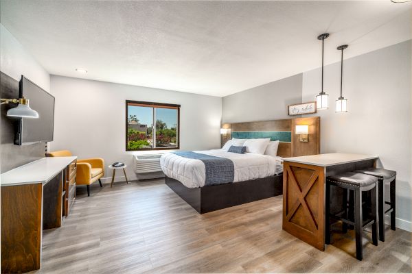 The image shows a modern hotel room with a king-sized bed, a wall-mounted TV, a desk, a chair, pendant lights, and a window with a view.