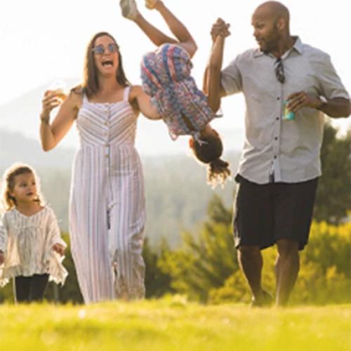 A family enjoys a sunny day outdoors, with parents swinging one child by their hands while another child happily walks alongside them.