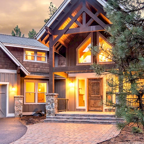 A large modern cabin-style house with warm glowing lights, surrounded by trees and featuring a three-car garage and a stone pathway.