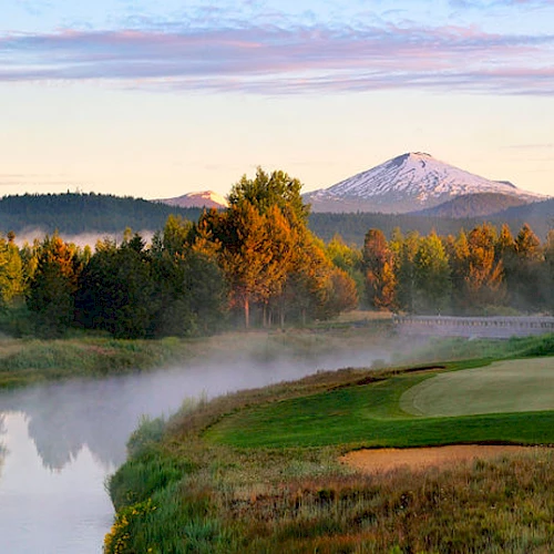 A tranquil golf course with a putting green by a serene river, surrounded by trees, and a snow-capped mountain under a sunrise sky in the background.