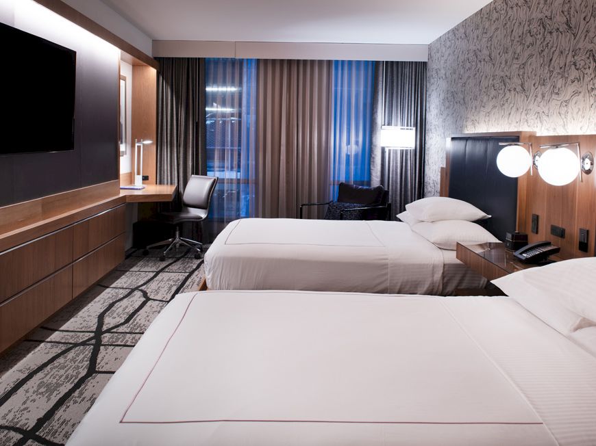 A modern hotel room with two beds, a wall-mounted TV, a desk with a chair, and patterned wallpaper behind the beds.