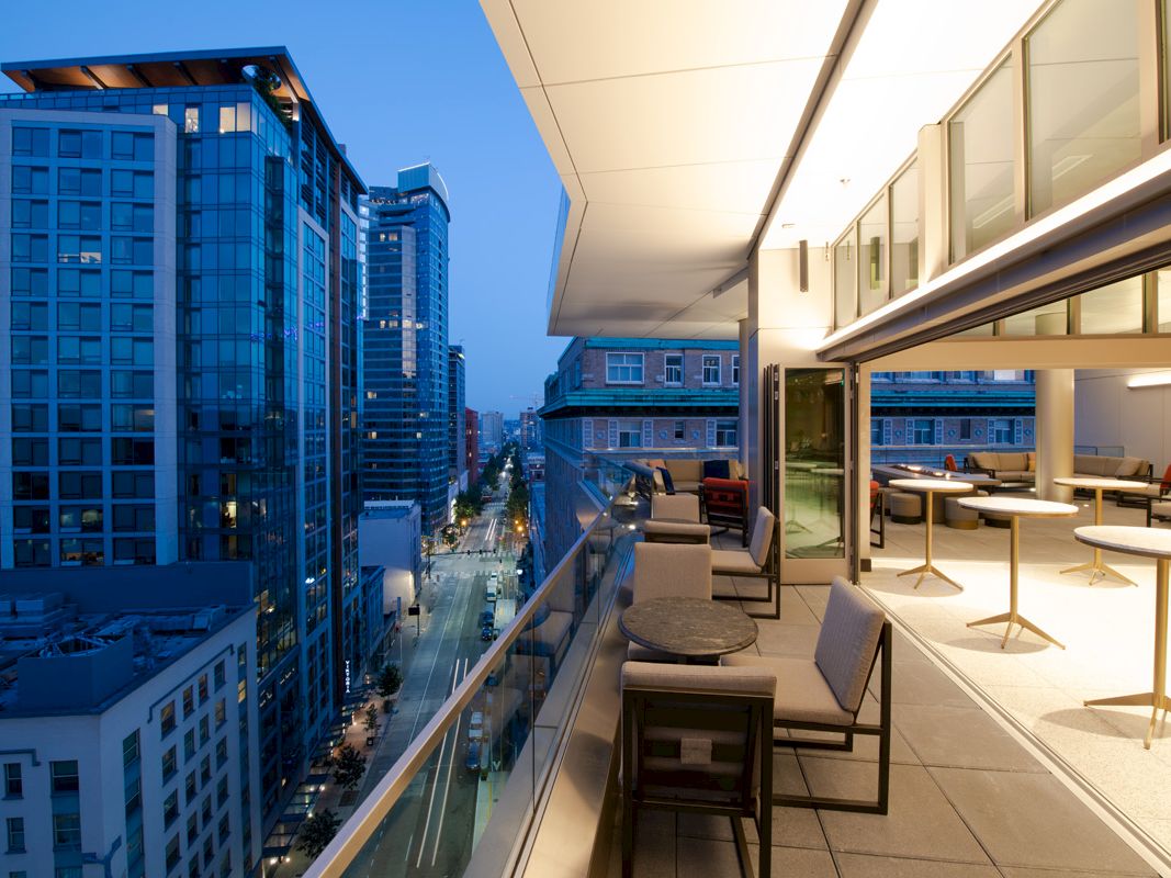 A modern cityscape at dusk with tall buildings and an illuminated, stylish outdoor balcony area featuring tables and chairs.