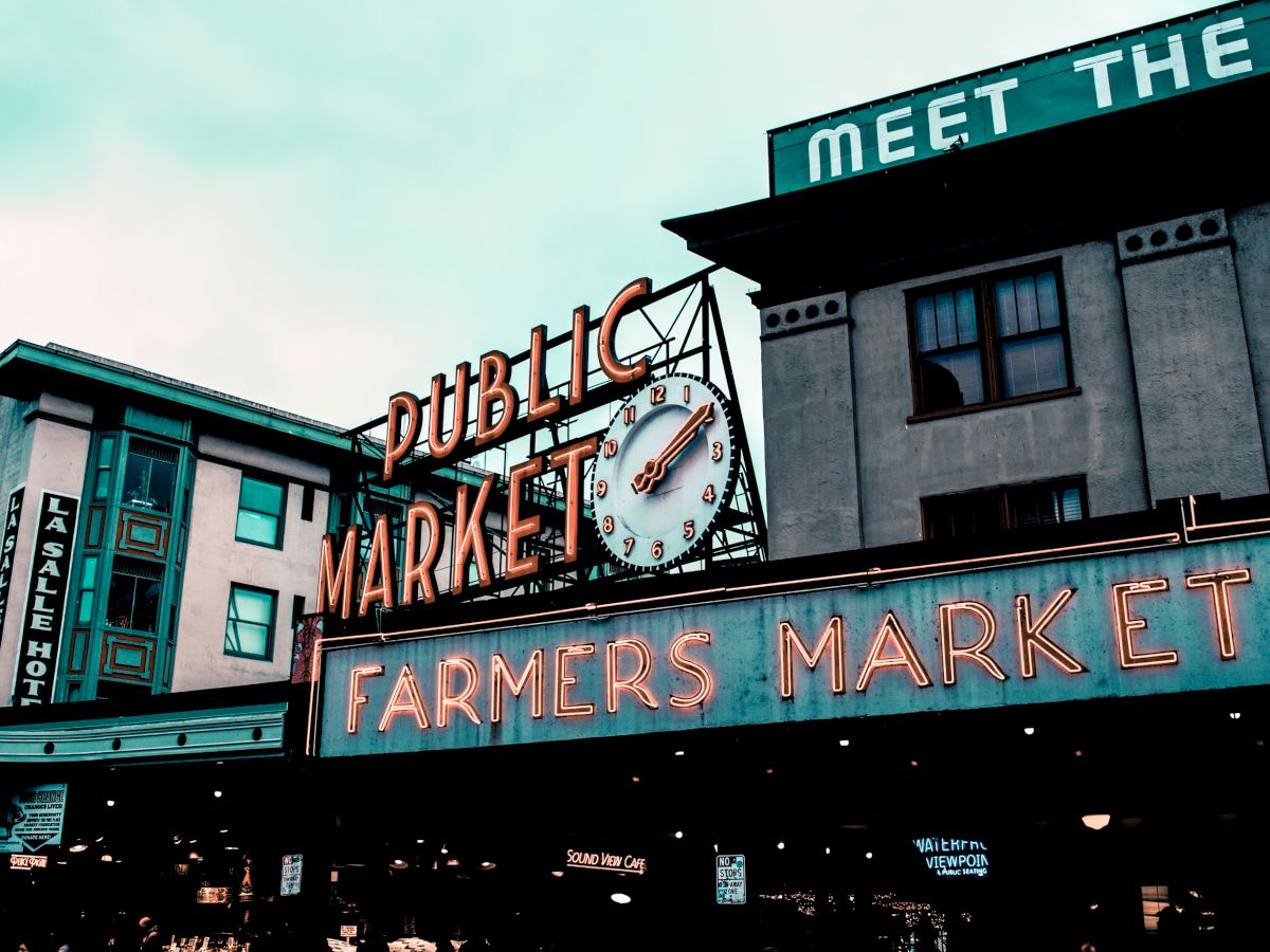 The image shows a vibrant public market sign with neon lights displaying "Farmers Market" in an urban area, featuring buildings in the background.