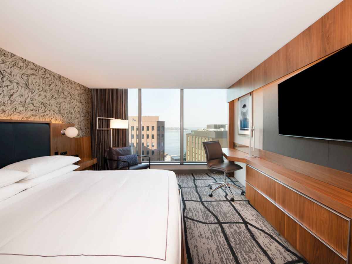 A modern hotel room features a large bed, wall-mounted TV, desk, chair, and a window with a view of buildings and a body of water.
