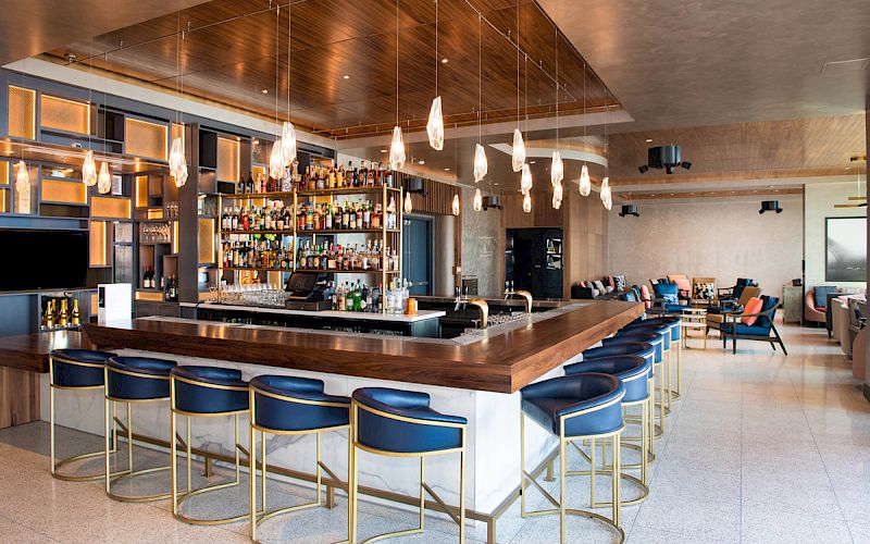 The image shows a modern, well-lit bar area with a wooden countertop, blue chairs, hanging lights, and a fully stocked bar situated centrally.