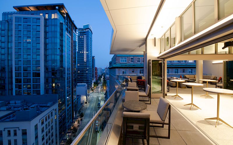 A modern rooftop balcony overlooks a cityscape with tall buildings at dusk, featuring outdoor seating and tables along the railing.