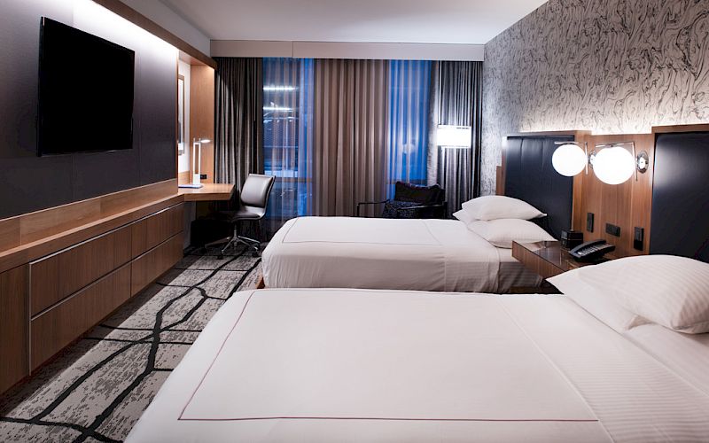 The image shows a modern hotel room with two neatly made double beds, a wall-mounted TV, a desk, and stylish lighting.