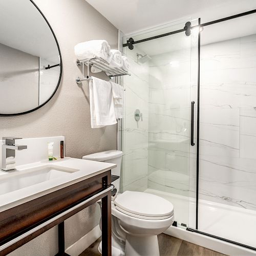 The image shows a modern bathroom with a round mirror, sink, toilet, and glass-enclosed shower. Towels are neatly stacked on a rack.