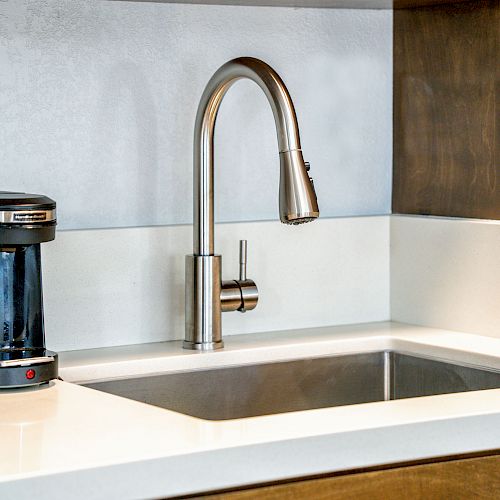 A modern kitchen counter with a stainless steel sink and a faucet, accompanied by a small coffee maker, all on a white countertop.