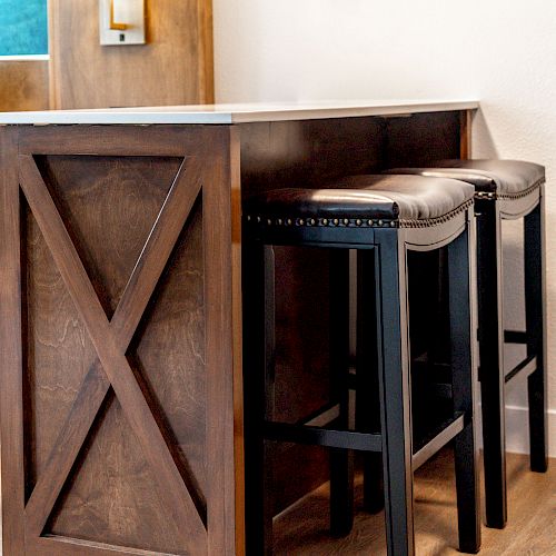 A small bar-height counter with a wooden X-frame design and two upholstered stools, placed beside a bed in a cozy room.