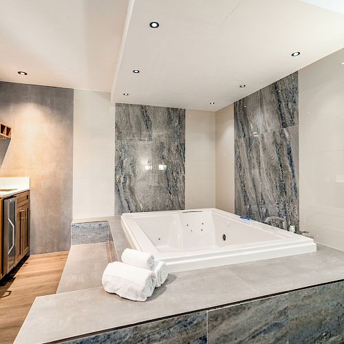 This image shows a modern bathroom with a large bathtub, marble walls, wooden cabinetry, and white towels on the edge of the tub.