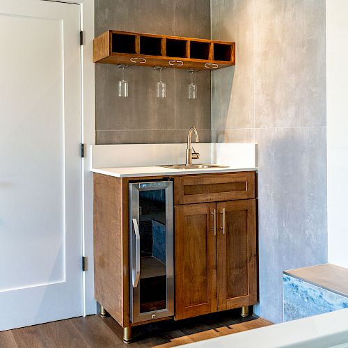 The image shows a small kitchenette area with a wooden cabinet, small sink, wine glass rack, mini-fridge, and a modern gray tile wall.