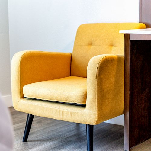 A yellow armchair with black legs is placed beside a wooden desk on a light wood floor, suggesting a cozy and stylish interior space.