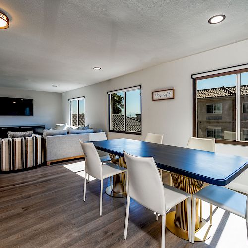 A modern open-concept dining and living area with a blue dining table, white chairs, striped armchairs, a couch, and large windows letting in natural light.